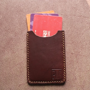 K&ouml;nnte beinhalten: Ein brauner Lederkartenhalter mit drei teilweise sichtbaren Karten. Der Kartenhalter hat einen gen&auml;hten Rand und die Initialen "P.U.G." sind eingepr&auml;gt. Die Karten sind rot, orange und lila. Ungef&auml;hr 10 cm hoch.