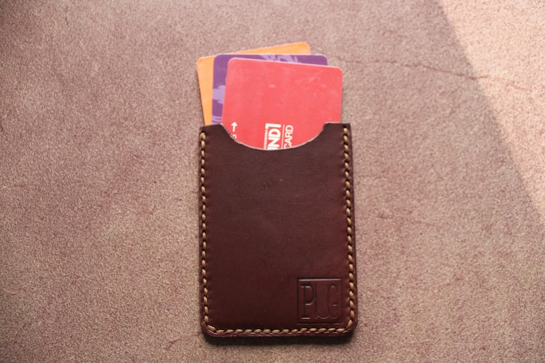 K&ouml;nnte beinhalten: Ein brauner Lederkartenhalter mit drei teilweise sichtbaren Karten. Der Kartenhalter hat einen gen&auml;hten Rand und die Initialen "P.U.G." sind eingepr&auml;gt. Die Karten sind rot, orange und lila. Ungef&auml;hr 10 cm hoch.