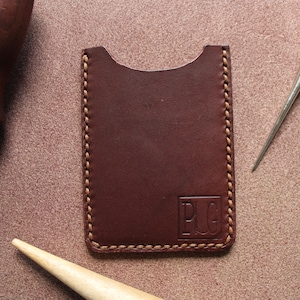 K&ouml;nnte beinhalten: Dunkelbrauner Lederkartenhalter mit gen&auml;hten Kanten und gepr&auml;gten Initialen "PLG". Das Bild zeigt auch Lederwerkzeuge, wie eine Ahle und einen Nahtformer, auf einem braunen Hintergrund.