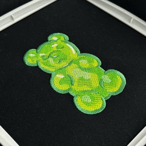May include: A green embroidered gummy bear patch with a light green outline and details, set against a black fabric background. The patch is in a white embroidery hoop.