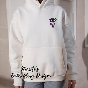 Op de afbeelding: Witte hoodie met een zwart en groen oog met tranen ontwerp. De tekst "Marite's Embroidery Designs" is gedrukt op de voorkant van de hoodie.