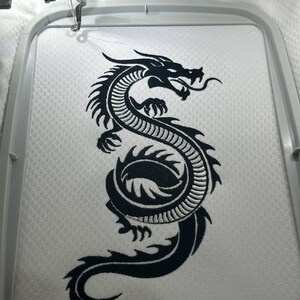 Puede incluir: Dise&ntilde;o de bordado de un drag&oacute;n negro sobre tela blanca, enmarcado por un bastidor de bordado. El drag&oacute;n est&aacute; detallado con escamas y una cola fluida, listo para un proyecto personalizado. La m&aacute;quina de bordar es visible en la parte superior de la imagen.
