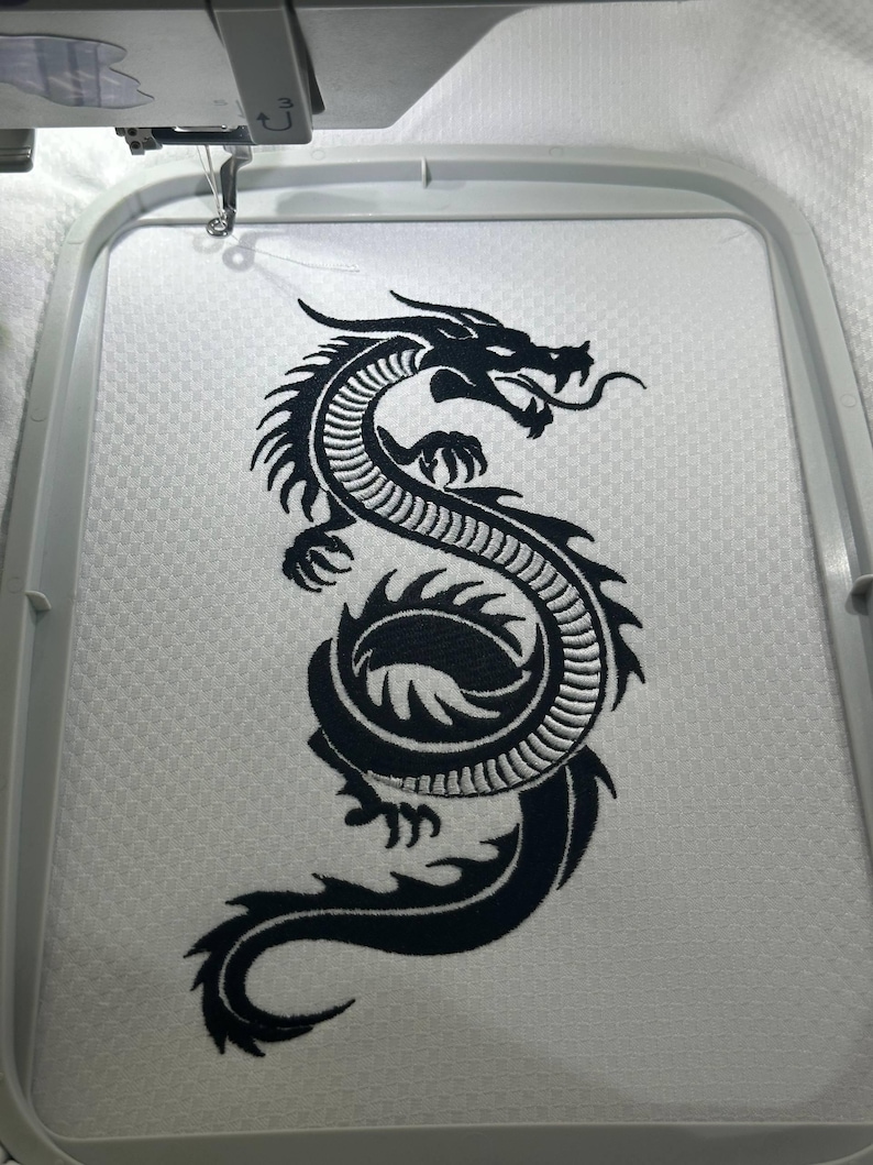Puede incluir: Dise&ntilde;o de bordado de un drag&oacute;n negro sobre tela blanca, enmarcado por un bastidor de bordado. El drag&oacute;n est&aacute; detallado con escamas y una cola fluida, listo para un proyecto personalizado. La m&aacute;quina de bordar es visible en la parte superior de la imagen.