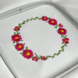 May include: Embroidered floral wreath design in a gray embroidery hoop. The wreath features vibrant pink flowers with yellow centers and green leaves, set against a white, textured fabric background. The embroidery is detailed and colorful.