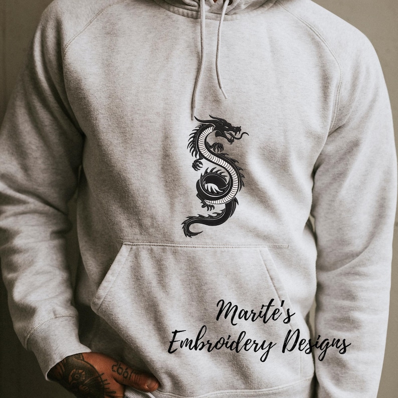 Puede incluir: Sudadera con capucha gris claro con un dise&ntilde;o de drag&oacute;n negro en la parte delantera. El drag&oacute;n est&aacute; estilizado y detallado. La sudadera tiene un bolsillo de canguro y una capucha con cord&oacute;n. El texto "Marite's Embroidery Designs" est&aacute; impreso en la parte inferior derecha.