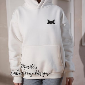 Puede incluir: Sudadera con capucha blanca con un gato negro asom&aacute;ndose desde detr&aacute;s del bolsillo. El texto "Marite's Embroidery Designs" est&aacute; impreso en la parte inferior de la sudadera.