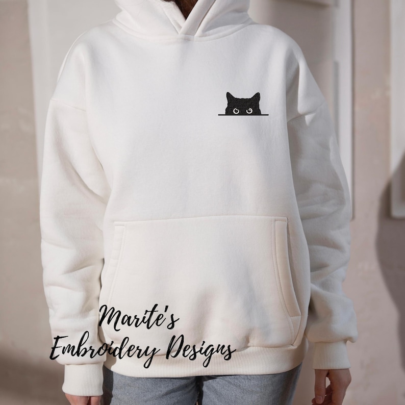 Puede incluir: Sudadera con capucha blanca con un gato negro asom&aacute;ndose desde detr&aacute;s del bolsillo. El texto "Marite's Embroidery Designs" est&aacute; impreso en la parte inferior de la sudadera.