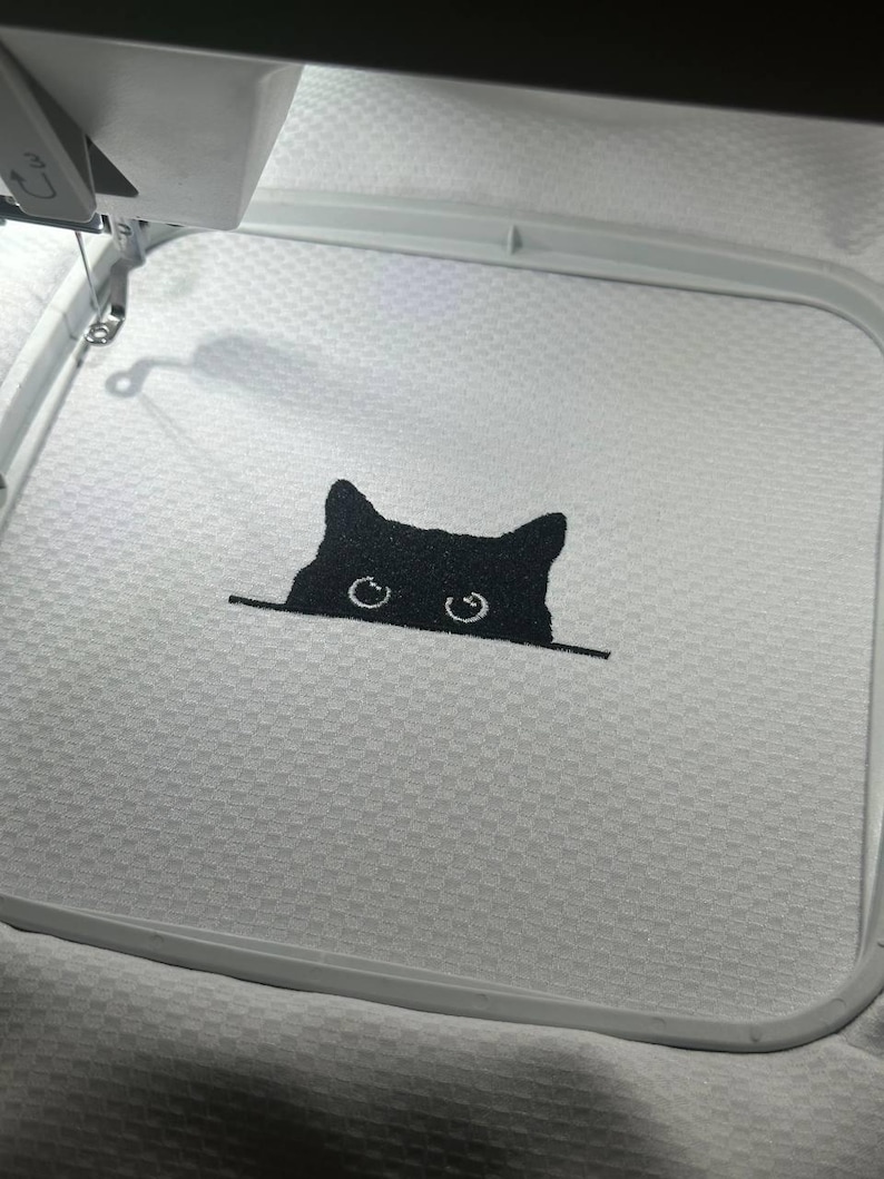 Puede incluir: Un gato negro asom&aacute;ndose desde detr&aacute;s de una superficie blanca. Los ojos del gato son grandes y redondos, y su pelaje es negro. El gato est&aacute; bordado en una tela blanca.