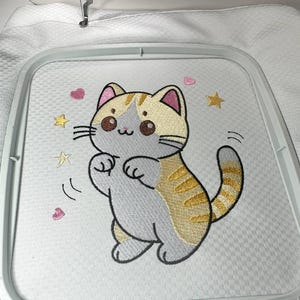 以下が含まれることがあります： ベージュとグレーの漫画の猫の刺繍デザイン。金色の星とピンクのハートに囲まれています。猫は遊び心のあるポーズで、刺繍枠の中で白い布にステッチされています。
