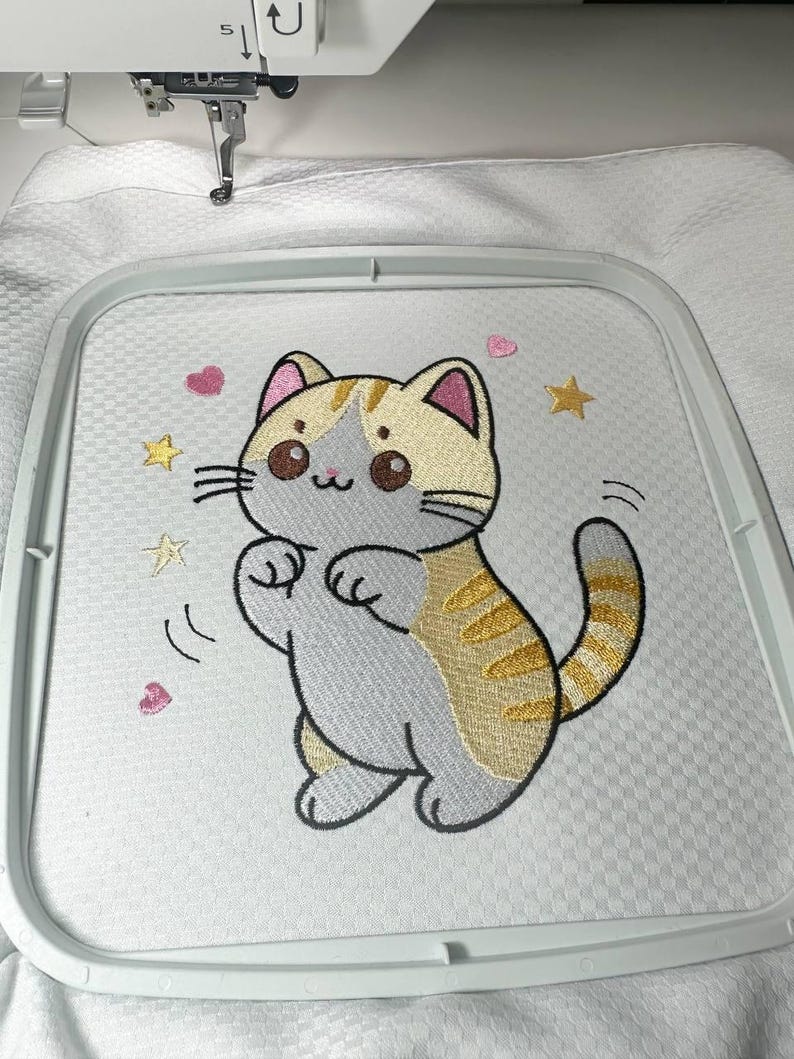 以下が含まれることがあります： ベージュとグレーの漫画の猫の刺繍デザイン。金色の星とピンクのハートに囲まれています。猫は遊び心のあるポーズで、刺繍枠の中で白い布にステッチされています。