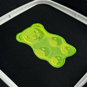 Könnte beinhalten: Ein grün-gelber, gestickter Gummibären-Aufnäher auf schwarzem Stoff, der in einem weißen Stickrahmen platziert ist. Das Gummibären-Design weist detaillierte Stickereien mit helleren gelben Akzenten auf.