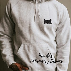Puede incluir: Una sudadera con capucha gris con un gato negro asom&aacute;ndose por detr&aacute;s de una l&iacute;nea blanca. El texto "Marite's Embroidery Designs" est&aacute; impreso en la parte inferior de la sudadera.