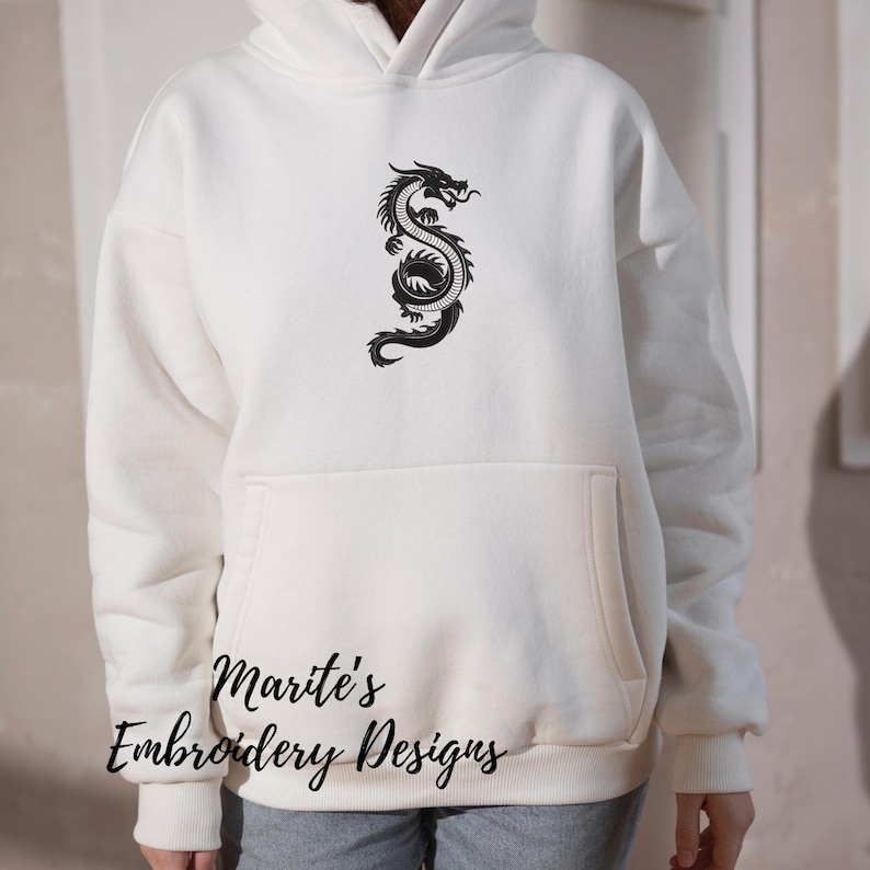 Puede incluir: Sudadera con capucha color crema con un dise&ntilde;o de drag&oacute;n negro en la parte delantera. La sudadera tiene un bolsillo de canguro y una capucha con cord&oacute;n. El texto "Marite's Embroidery Designs" est&aacute; impreso en la parte inferior.