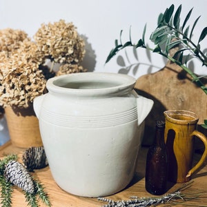 May include: A cream-colored, antique-style ceramic pot with two handles and horizontal etched lines. The pot is displayed with dried flowers, a brown bottle, and a yellow pitcher on a wooden surface.