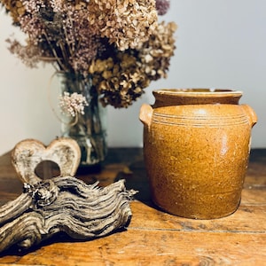 May include: A rustic brown ceramic crock with a wide mouth and two handles. The crock is sitting on a wooden table with a piece of driftwood and dried flowers in the background.