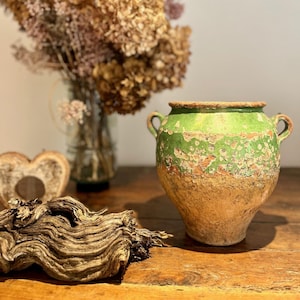 May include: A green glazed ceramic pot with a rustic, aged look. The pot has a wide, round body and two handles. The pot is sitting on a wooden surface.