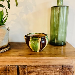 Puede incluir: Un macetero de cerámica rayado en verde, amarillo y marrón con dos asas. El macetero está sobre una superficie de madera.