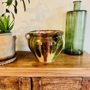 May include: A large, green, brown, and yellow glazed ceramic pot with two handles. The pot is sitting on a wooden table next to a green glass bottle.