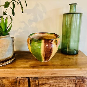 Op de afbeelding: Een grote, groene, gele en bruine geglazuurde keramische pot met twee handvatten. De pot staat op een houten oppervlak.