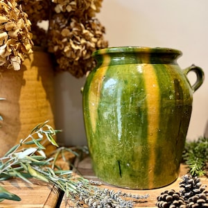 May include: A green and yellow striped ceramic pot with two small handles. Dried flowers and greenery are arranged around the pot on a wooden surface. The pot has a rustic, aged appearance.