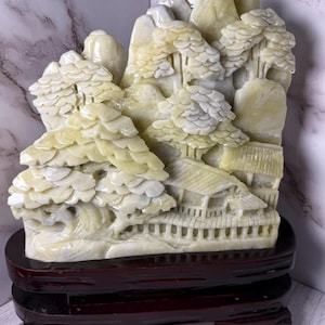 May include: A carved stone sculpture depicting a mountain landscape with trees and buildings. The sculpture is in shades of cream and light yellow, mounted on a dark brown wooden base. The intricate details include textured trees and architectural elements.