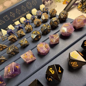 May include: A collection of polyhedral dice sets in various colors and materials, including black, purple, and gray. The dice are displayed on a blue surface, with some sets labeled "Polyhedral Dice Set". The dice feature gold numbering.