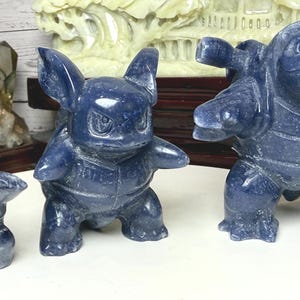 May include: Three dark blue stone carvings depicting the evolution of a turtle-like creature. The figures show a small rounded form, a medium-sized bipedal character with pointed ears, and a large bipedal figure with two cannons on its back.
