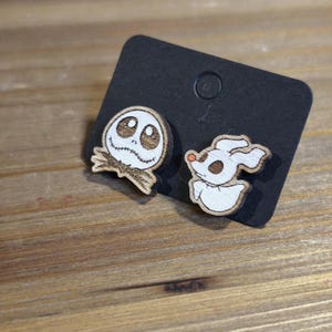 Può includere: Un paio di orecchini in legno con Jack Skellington e Zero di The Nightmare Before Christmas. Gli orecchini sono su una carta nera.