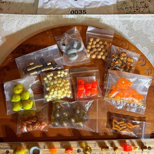 Op de afbeelding: Assortiment van kleurrijke kralen in kleine plastic zakjes. De zakjes zijn gerangschikt op een houten oppervlak. De kralen zijn in verschillende kleuren, waaronder oranje, geel, bruin, wit en zwart. De zakjes zijn gelabeld met nummers.
