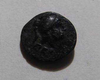 May include: A dark, circular, metallic ancient coin featuring a raised profile of a figure wearing a helmet facing toward the right. The surface of the coin shows signs of age, oxidation, and wear, with a textured, uneven patina across the metal.