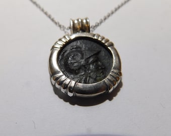 May include: A silver pendant necklace featuring a dark, circular ancient coin set within a polished silver frame. The coin displays a profile relief of a figure wearing a helmet. The pendant hangs from a thin silver chain, showcasing a metallic finish.