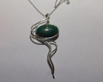 May include: A silver pendant necklace featuring an oval-shaped dark green stone with brown veining. The stone is set within a polished silver frame that extends into a series of flowing, curved silver wire lines that taper downward.