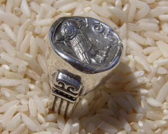 May include: A silver-toned ring featuring a circular face with an embossed owl design and Greek lettering. The band of the ring displays a decorative pattern with vertical lines and scrollwork details on the sides of the metal setting.