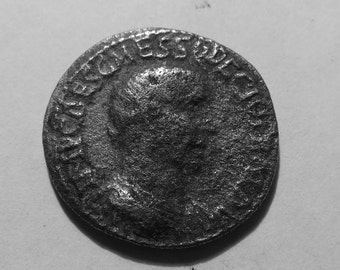 May include: A circular, dark-toned metallic coin featuring a profile portrait of a figure facing right. The surface has a weathered, textured appearance with raised lettering along the outer edge, showing signs of age and oxidation throughout.