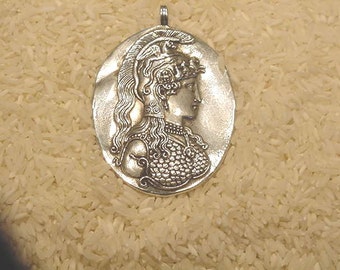 May include: A silver-colored pendant with a detailed profile of a woman in classical attire. The oval-shaped pendant features a raised relief design and a small loop at the top for a chain. The background is a bed of white rice.