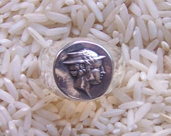 May include: A silver ring featuring a circular face with a relief profile of a figure wearing a winged helmet. The metal has a polished finish with darkened recessed areas that highlight the detailed features of the embossed classical design.