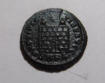 May include: A dark, circular, metallic coin featuring a central relief of a rectangular building with a brick-like pattern and a small arched doorway. The coin has a textured surface, a visible crack near the top edge, and faint lettering along the rim.
