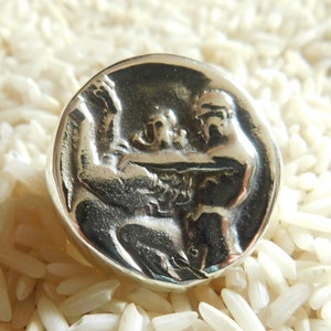 May include: A silver ring with a detailed design of two figures holding hands. The ring is set against a background of white rice.