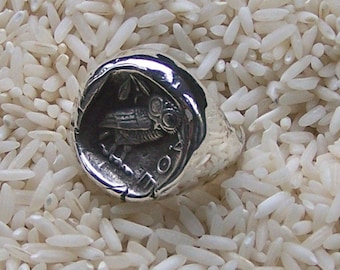 May include: A silver ring with an owl design. The owl is facing right and has its wings spread. The ring is set against a background of white rice.