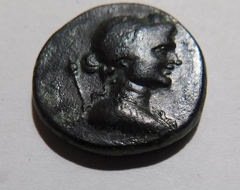 May include: A dark, circular metallic coin featuring a raised profile of a person facing right. The figure has hair pulled back and wears a garment with visible texture. A thin, vertical staff-like element is positioned behind the head.