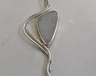 May include: A silver-toned pendant necklace featuring a triangular, light gray druzy stone set within a polished metal frame. The setting extends into a fluid, asymmetrical, wavy metal design that curves alongside the stone, suspended from a chain.