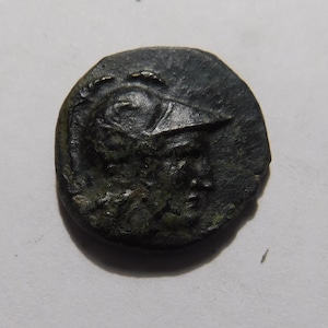 May include: A dark, aged coin featuring a detailed relief of a helmeted figure. The coin's surface shows signs of wear and age. The design suggests a historical or ancient artifact, possibly Roman or Greek.