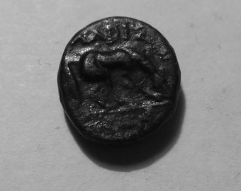 May include: A dark, circular, metallic coin featuring a raised relief of a four-legged animal in profile. The surface shows signs of age and wear with a textured, uneven patina. Above the animal, there are faint, partially visible characters.