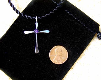 Puede incluir: Un colgante de cruz de plata con una piedra preciosa púrpura en el centro, colgando de un collar de cordón negro. El collar se muestra en una bolsa de terciopelo negro.
