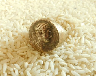 May include: A metallic ring featuring a circular face with a raised relief profile of a person in a classical style. The ring has a weathered, aged finish with a muted gold and bronze tone, showcasing a detailed portrait embossed on the front surface.