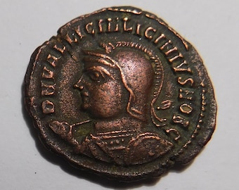 May include: A circular bronze-toned ancient coin featuring a profile relief of a figure wearing a crested helmet. The coin surface displays raised lettering around the perimeter that reads DNVALILICINIVSNOBC, with a textured, weathered patina.