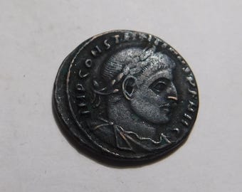 May include: A dark, circular ancient bronze coin featuring a profile portrait of a ruler wearing a laurel wreath. The text IMP CONSTANTINVS AVG is embossed along the outer edge of the coin, surrounding the central relief of the head.