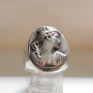 May include: A silver ring with a raised image of a woman's profile wearing a crown. The ring is oval shaped and has a smooth, polished finish.