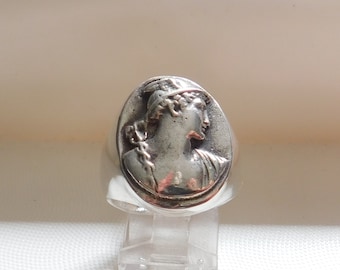 May include: A silver ring with a raised image of a woman's profile wearing a crown. The ring is oval shaped and has a smooth, polished finish.
