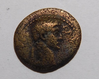 May include: A circular bronze-toned ancient coin featuring a weathered profile portrait of a figure facing right. The surface shows a textured, pitted patina with dark brown and metallic gold highlights, and a beaded border along the outer edge.